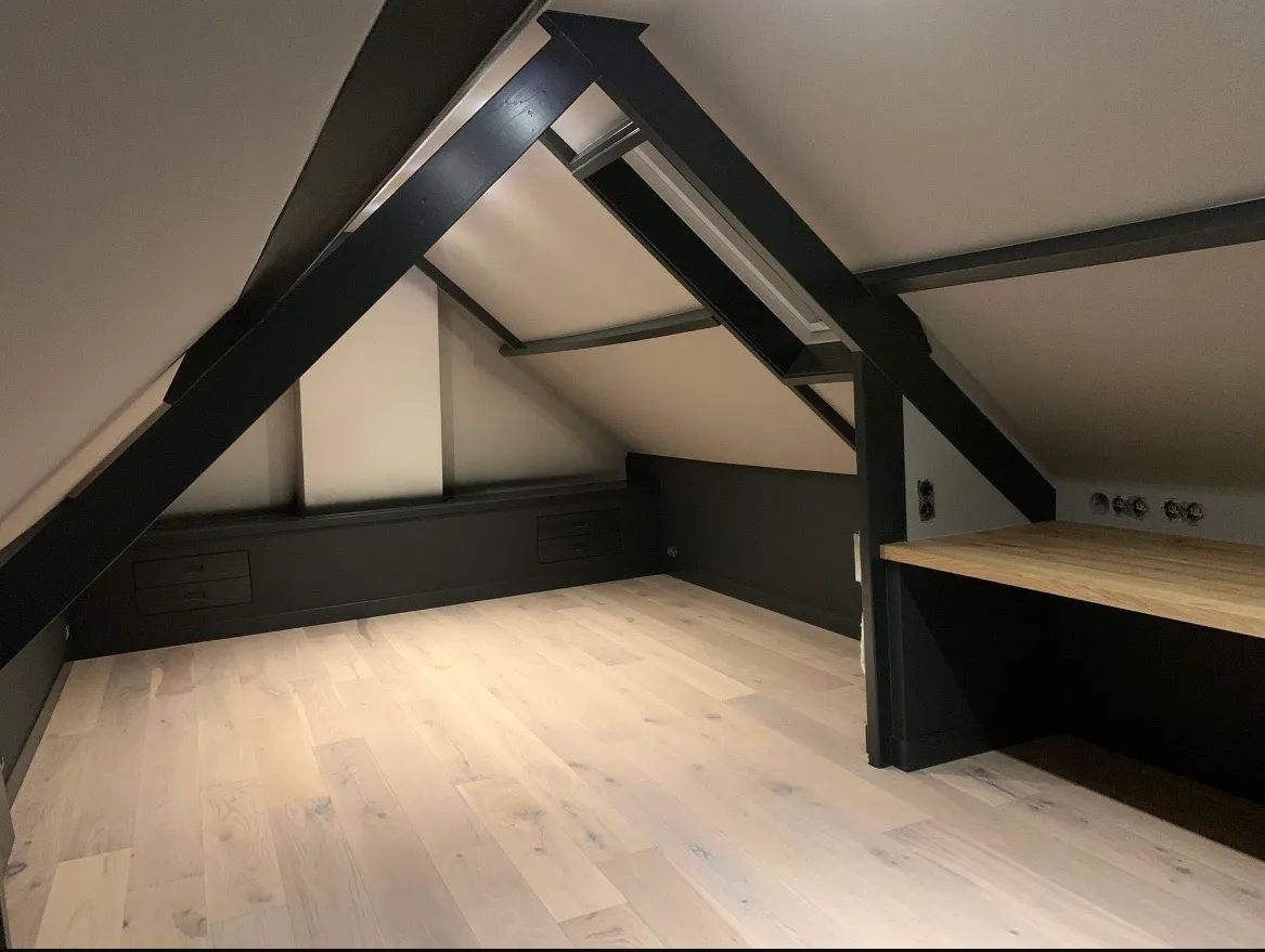 Zolderkamer met donker geschilderde spanten en kozijnen boven een lichte houten vloer.