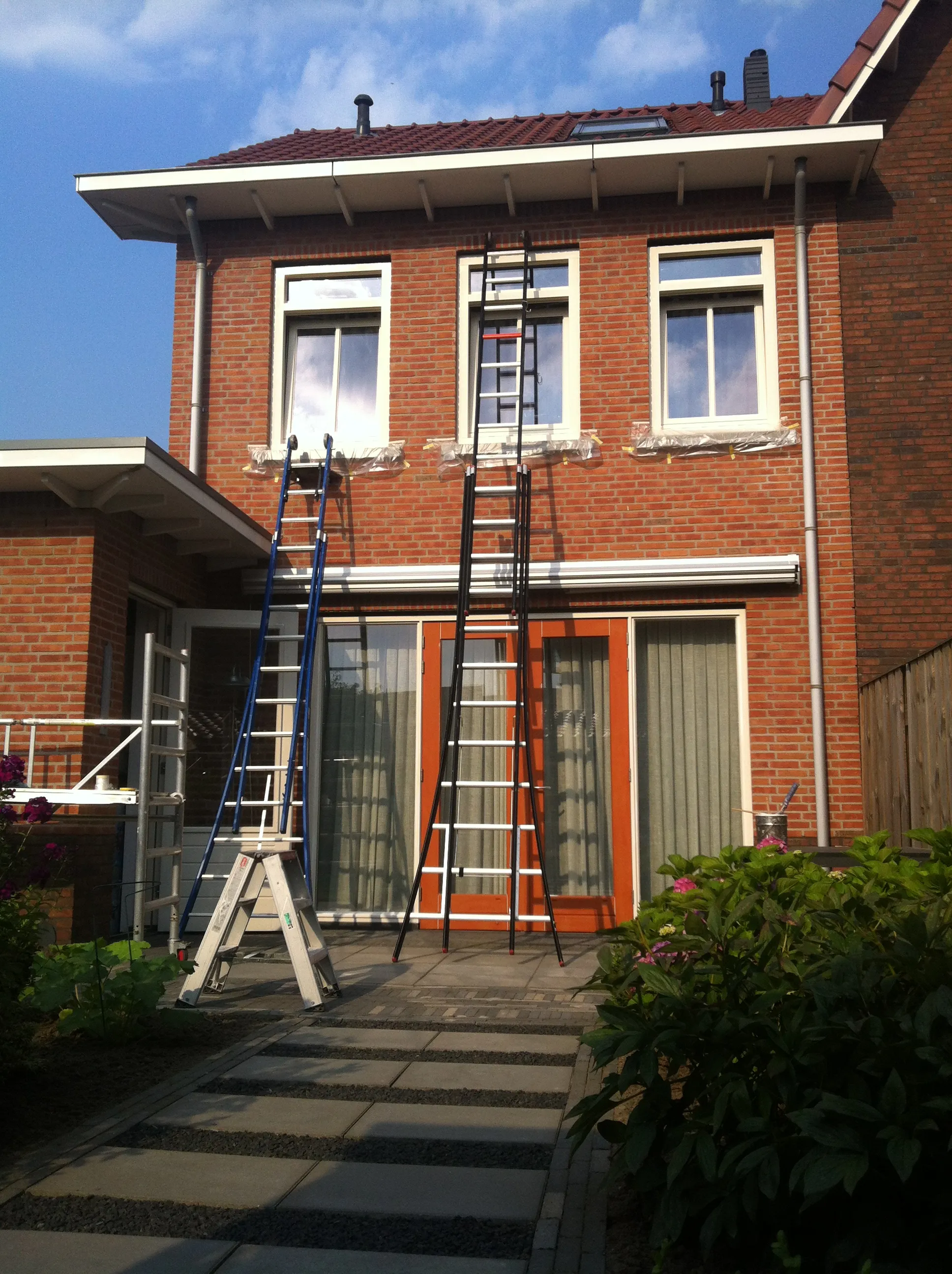 Buitenschilderwerk in uitvoering: ladders tegen de bakstenen gevel van een woonhuis, de kozijnen afgeplakt en in de grondverf.